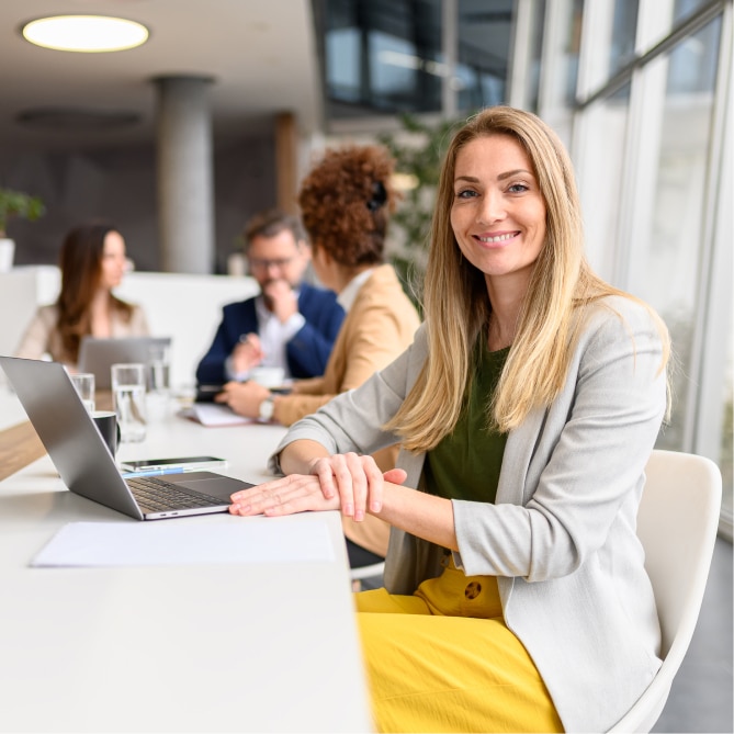 A smiling woman with blonde hair sits at a white office table wearing a grey blazer and yellow trousers. She is looking toward the camera with a laptop open in front of her. In the blurred background, several colleagues are seated at the same long table in a bright, modern office space.